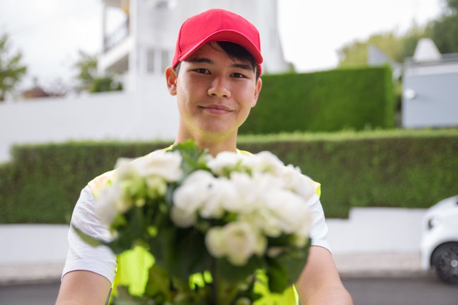 A smiling young male flower delivery courier wearing a red cap and a high-visibility yellow vest, handing over a lush bouquet of white roses and greenery to a recipient outdoors in an urban residentia