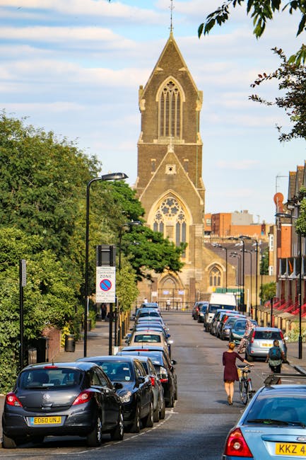A row of parked cars lining a gentle incline street in Lea Bridge, with a pedestrian and cyclist visible on the pavement. In the background, a historic church with a tall, pointed spire and intricate