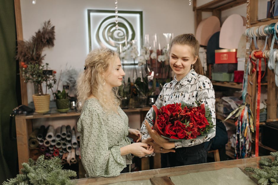 A young woman in a floral blouse is holding a large, vibrant bouquet of fresh red roses, adorned with greenery and small red accent flowers, wrapped partially in green paper. She stands behind a woode