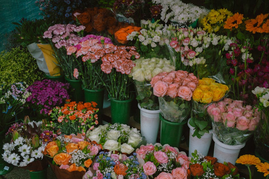 A display of various fresh flower bouquets arranged in white and green buckets at a flower shop, featuring roses, carnations, daisies, chrysanthemums, and other blooms in colors such as pink, white, y