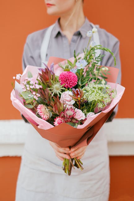 A person dressed in a light grey shirt and white apron holds a large bouquet of fresh flowers, wrapped in pink and brown paper, in front of a warm orange background. The bouquet features a variety of