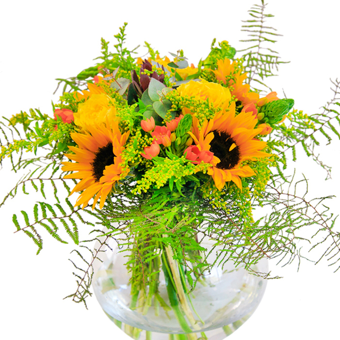 Sunlit exuberance radiates from this abundant arrangement, thoughtfully gathered by a Leabridge florist with an eye for community-inspired beauty. The bouquet unfurls with a joyful profusion of sunflowers-each vivid yellow petal and bold, brown center reminiscent of late summer fields stretching across Hackney Marshes. Spiraled yellow roses nestle beside them, their petal layers rich and plush, catching and reflecting soft morning light so familiar to those living near Clapton Park. Sprigs of scarlet hypericum berries, their glossy sheen unmistakable, glimmer like tiny lanterns among the bouquet's greenery, creating striking accents that heighten the vibrancy of the dominating yellows and lush greens. Feathery ferns cascade gently around the arrangement's edges, their wispy tips adding kinetic energy and a sense of wild, garden-inspired movement. Delicate spikes of verdant filler-likely solidago-stitched with minute yellow blossoms, alongside silver-tinged eucalyptus leaves, add texture and a touch of countryside nostalgia. All these blossoms are cradled in a clear, rounded glass vase, allowing the water to glimmer and the stems to create elegant, natural lines. This bouquet, brimming with the freshness and warmth of Leabridge, is a perfect tribute for joyful milestones or to enliven any setting, reflecting the uplifting spirit found along Chatsworth Road and crafted with care by Flower Delivery Lea Bridge.