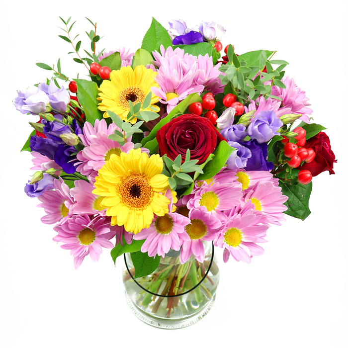 In this image, a tall, cylindrical glass vase holds a lavish mixed bouquet that seems to billow outward, set before a bright white backdrop that highlights every colour and texture. Deep red roses form the emotional anchor at the centre, their plush, velvety petals opening in generous spirals that hint at timeless love and appreciation. Framing them are two striking yellow gerbera daisies, each with a pronounced dark brown-orange disc at its heart, like miniature suns warming the whole display. Around these bold shapes, soft pink daisies and chrysanthemums spread in gentle waves, their countless slender petals and tiny yellow centres giving the arrangement a comforting, almost nostalgic charm. Threaded elegantly among the pinks are purple and lavender lisianthus, their bell-like blooms adding cool sophistication, as if echoing the serene tones of the River Lea at dusk. Small, spherical red hypericum berries are dotted throughout, glossy and bright, contributing a slightly playful note and textural interest. Abundant greenery-broad dark leaves, lighter lime-toned stems, and airy filler foliage-supports and surrounds the flowers, making the bouquet appear lush and freshly gathered. The stems are clearly visible through the transparent vase, neatly trimmed and fanned in clear water, a detail that speaks to the skilled hand of a local florist familiar with Lea Bridge homes and celebrations. The overall impression is of a joyful, celebratory arrangement, ideal for birthdays, congratulations, or simply to bring a burst of colour to a dining table near Lea Bridge Road, radiating warmth and natural beauty into the space.