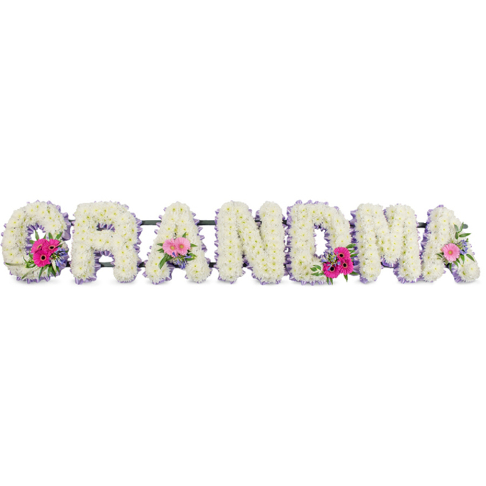 In this photograph, the word GRANDMA stretches horizontally across the frame as a floral tribute designed for a funeral or memorial. Each letter is built on a sturdy white base and lavishly covered with tight clusters of white chrysanthemums, placed so close together there is hardly any space between them. The blossoms look soft and cushiony, suggesting comfort and a sense of being held. Around the outer edge of every letter runs a narrow border of lilac ribbon, folded and pleated with care. This ribbon edging subtly lifts the crisp white blooms, adding a gentle wash of colour that feels soothing rather than showy, much like a soft lilac scarf worn on a cool Lea Bridge morning. At three points, small posy-style arrangements are nestled into the letters: vibrant pink gerberas fan out their daisy-like petals, mingling with deeper and paler pink roses, some fully open, others still cupped in bud. Fresh green foliage is threaded through these mini bouquets, providing contrast and a hint of natural movement, as if a breeze from nearby Lea Bridge Road or the River Lea had just brushed across the flowers. The lighting is even and calm, avoiding harsh glare so the layers of texture - the ruffled petals, the smooth satin ribbon, the glossy leaves - can be clearly seen. This arrangement feels deeply personal, the kind of piece a family might choose when saying goodbye in a local chapel or crematorium, created to stand quietly beside the coffin and speak of gratitude, gentleness, and many years of shared love.